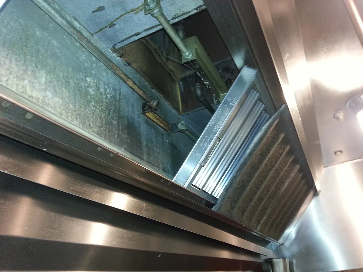 Close-up looking up into a stainless commercial exhaust hood, showing installed filter panels and the grease channel